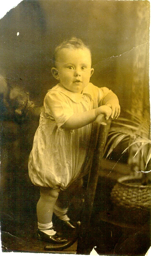 Original sepia photograph of a baby standing by a chair, with creases and damage